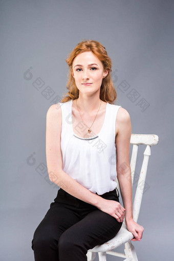 美丽的年轻的高加索人女人长红色的头发高高跟鞋坐着椅子黑色的裤子白色衬衫完整的增长灰色的背景业务概念