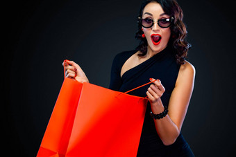 黑色的星期五出售概念商店惊讶女人太阳镜持有红色的袋孤立的黑暗背景购物