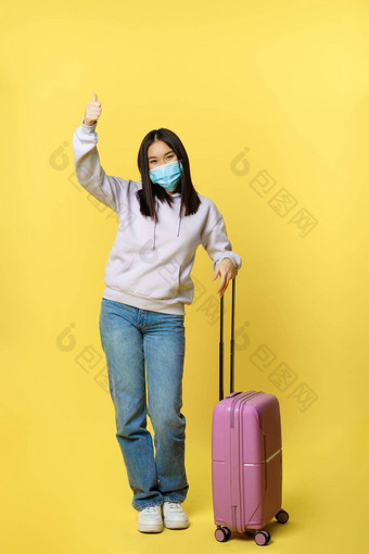 完整的长度拍摄热情的朝鲜文女孩享受假期摆姿势手提箱穿脸医疗面具旅行在国外科维德流感大流行黄色的背景