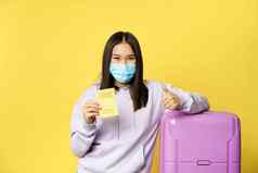 微笑亚洲女孩旅游脸面具站手提箱显示科维德国际疫苗接种证书旅行者拇指黄色的背景