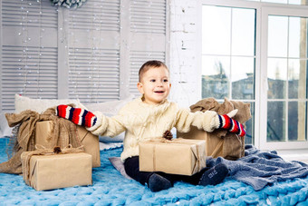 有趣的好玩的男孩孩子坐在床上圣诞节一天礼物盒子白色羊毛针织毛衣大明亮的连指手套笑着说大声室内节日装饰