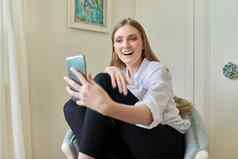 年轻的金发女郎坐着首页椅子视频沟通智能手机