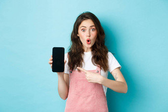 深刻的印象年轻的现代女人指出智能<strong>手机屏幕</strong>哇站在惊讶相机显示太棒了应用程序在线商店站蓝色的背景