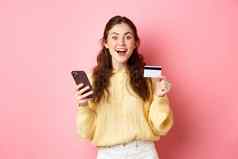技术在线购物兴奋女孩使订单支付在线塑料信贷卡持有移动电话微笑相机粉红色的背景