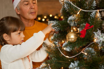 快乐家庭装修圣诞节树祖父穿橙色毛衣女孩白色跳投摆姿势生活房间美丽的圣诞节树泡沫弓