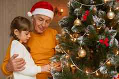 快乐家庭摆姿势室内圣诞节树祖父橙色衬衫圣诞老人老人他拥抱迷人的（外）孙女装修冷杉树庆祝一年