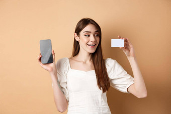 年轻的高加索人女人显示塑料信贷卡很高兴微笑<strong>演示</strong>空<strong>手机</strong>屏幕站米色背景