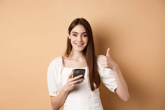 在线购物概念微笑快乐女人显示拇指智能手机持有电话站米色背景