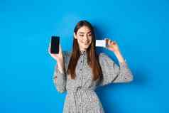 在线购物美丽的女模型显示空手机屏幕塑料信贷卡微笑很高兴购买互联网商店蓝色的背景