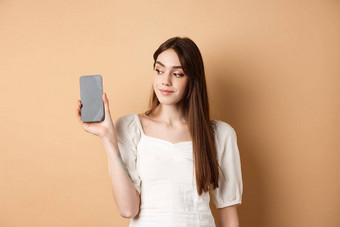 有吸引力的年轻的女人显示空智能手机屏幕微笑站米色背景