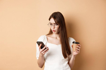 震惊了年轻的女人阅读移动电话屏幕皱着眉头持有咖啡杯站米色背景