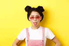 夏天时尚概念愚蠢的亚洲青少年女孩显示舌头穿太阳镜站黄色的背景