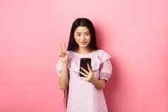 在线购物时尚的亚洲青少年女孩智能手机显示和平标志微笑很高兴相机站粉红色的背景