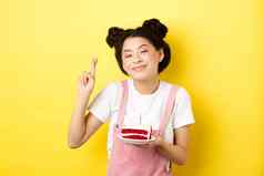 假期庆祝活动积极的亚洲生日女孩交叉手指使生日蛋糕基斯蜡烛微笑快乐相机黄色的背景