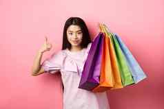 购物时尚的女孩显示色彩斑斓的纸袋竖起大拇指推荐商店折扣站粉红色的背景