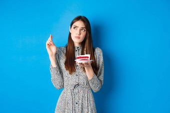 深思熟虑的生日女孩使交叉手指好运气持有蛋糕上左角落里蓝色的背景