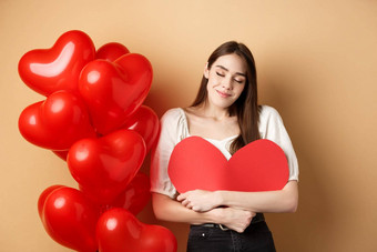 浪漫的女人拥抱大红色的心微笑梦幻下降爱情人节一天做梦情人站米色背景气球