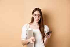 微笑年轻的女人眼镜阅读令人兴奋的新闻移动电话持有移动PC快乐相机站米色背景