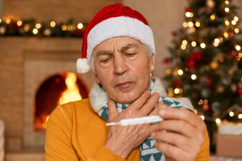 生病的高级男人。圣诞老人他坐着壁炉包装围巾穿圣诞老人老人他持有温度计手发热遭受痛喉咙提出了房间圣诞节装饰