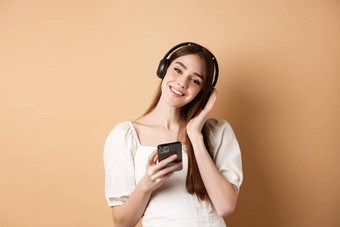 可爱的微笑女孩听音乐无线耳机智能手机站米色背景