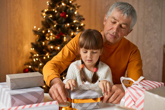 祖父（外）孙女包装礼物坐着表格房间圣诞节树背景孩子高级男人。穿休闲服装关系丝带弓