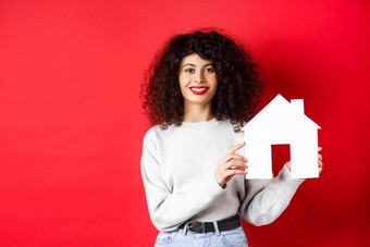 真正的房地产微笑高加索人女人卷曲的头发红色的嘴唇显示纸房子模型搜索财产站红色的背景