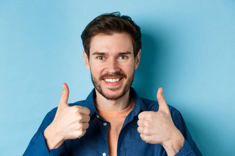 特写镜头微笑好看的男人。显示拇指赞扬好工作推荐公司站蓝色的背景