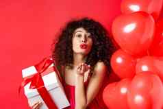 浪漫情人节一天概念漂亮的卷发女孩红色的衣服发送爱吹空气吻相机持有礼物情人站心红色的背景