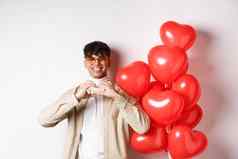 情人节一天浪漫概念英俊的年轻的男人。站红色的心气球显示心手势爱微笑白色背景