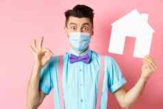 科维德流感大流行真正的房地产概念兴奋年轻的男人。医疗面具显示标志纸房子断路推荐机构站粉红色的背景