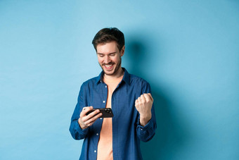快乐年轻的男人。赢得视频游戏移动电话满意智能<strong>手机屏幕</strong>快乐站蓝色的背景