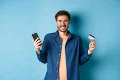 电子商务概念兴奋年轻的男人。持有智能手机塑料信贷卡购物在线站蓝色的背景