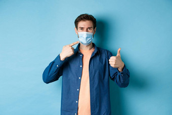 冠状病毒流感大流行概念年轻的健康的男人。指出医疗面具显示拇指<strong>预防措施</strong>捕捉科维德蓝色的背景