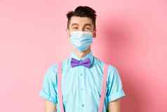 科维德流感大流行健康概念肖像微笑男人。医疗面具感觉快乐站节日蝴蝶结背带裤粉红色的背景