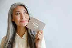 多梦的灰色头发的亚洲女人持有礼物卡脸工作室
