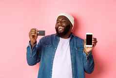 在线购物快乐非裔美国人男人。无檐小便帽笑显示信贷卡移动电话屏幕站粉红色的背景