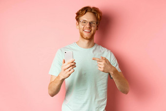 年轻的男人。红色的头发胡子穿t恤眼镜微笑指出手指智能手机推荐在线促销活动粉红色的背景