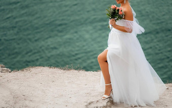 华丽的金发女郎新娘奢侈品白色衣服婚礼花束摆姿势岩石海岸平静海日落婚礼概念一边视图