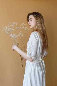 美丽的瘦年轻的女孩长头发白色衣服米色背景持有白色花手
