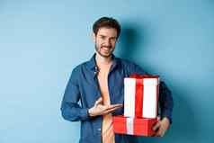 年轻的男人。休闲衣服购买浪漫的礼物情人节一天指出礼物盒子微笑站蓝色的背景