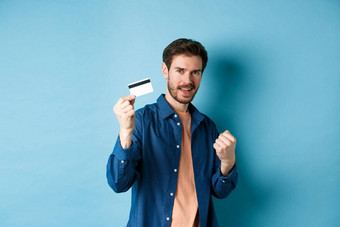 满意年轻的男人。显示塑料信贷卡欢呼快乐站蓝色的背景