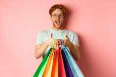 快乐的红色头发的人男人。购买礼物持有购物袋微笑站粉红色的背景