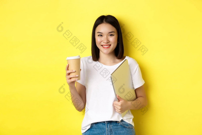 亚洲女孩学生喝咖啡持有移动PC微笑相机站黄色的背景