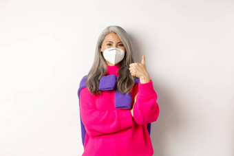 科维德流感大流行社会距离概念快乐的时尚的亚洲高级女人穿呼吸器冠状病毒显示竖起大拇指站白色背景