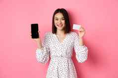 可爱的亚洲女人购物在线显示银行信贷卡移动屏幕微笑相机站粉红色的背景