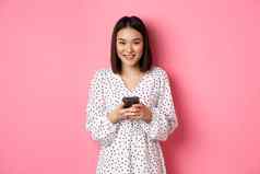 美丽的亚洲夫人阅读消息微笑移动电话站可爱的衣服粉红色的背景