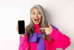在线购物美丽的亚洲祖母微笑指出手指智能手机空白屏幕惊讶显示移动应用程序站白色背景