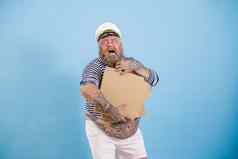 快乐有胡子的肥胖男人。水手持有空白盒子披萨摆姿势光蓝色的背景