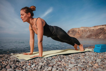 年轻的女人长头发健身教练黑色的运动服装紧身裤上衣伸展运动普拉提瑜伽席海阳光明媚的一天女健身瑜伽例程概念