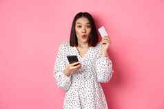 有吸引力的年轻的亚洲女人订单在线持有信贷卡移动电话使互联网购买站快乐粉红色的背景